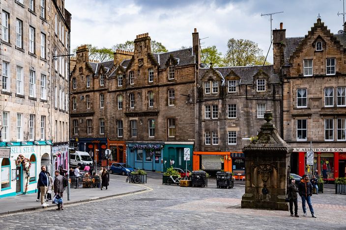A cobbled Edinburgh square edged with tall grey stone tenements and colourful ground-floor shopfronts; people stroll past a carved stone monument under a cloudy sky