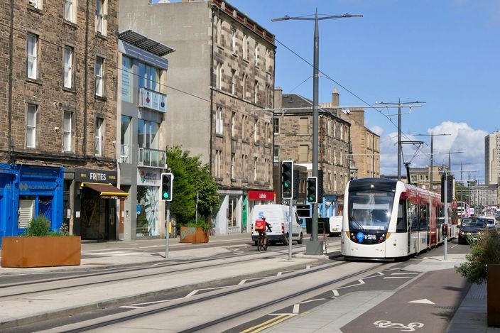 A white, black and red Edinburgh tram on street tracks running down Leith Walk, flanked by tenement buildings with shopfronts under a blue sky