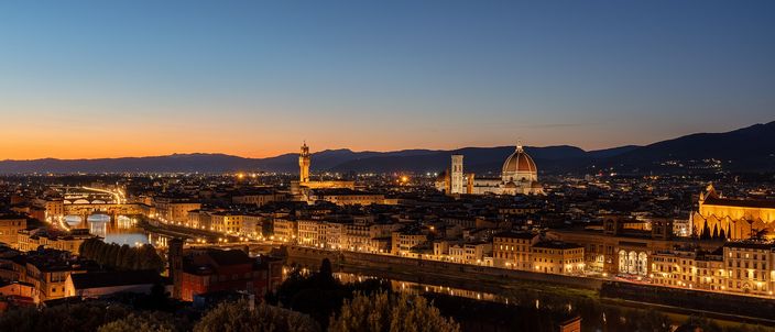 Florence at dusk seen from Piazzale Michelangelo: the lit dome of the cathedral and the tower of Palazzo Vecchio above the rooftops, with the Arno and its bridges in the foreground and the hills behind