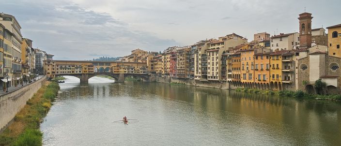 The Arno seen from a bridge, with the Ponte Vecchio and its overhanging shops spanning the river ahead, ochre and yellow houses lining the far bank, and a single sculler on the water
