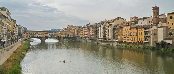 The Arno seen from a bridge, with the Ponte Vecchio and its overhanging shops spanning the river ahead, ochre and yellow houses lining the far bank, and a single sculler on the water