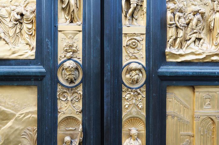 The two leaves of the Florence Baptistery's east door meeting at the centre: large square gilded bronze relief panels of biblical scenes set into a dark frame, with small gilt statuettes standing in niches down the central strip and portrait heads in round medallions between them