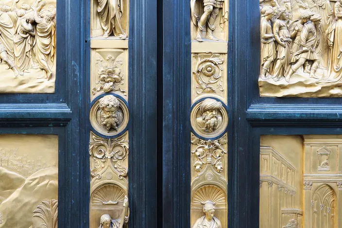 The two leaves of the Florence Baptistery's east door meeting at the center: large square gilded bronze relief panels of biblical scenes set into a dark frame, with small gilt statuettes standing in niches down the central strip and portrait heads in round medallions between them