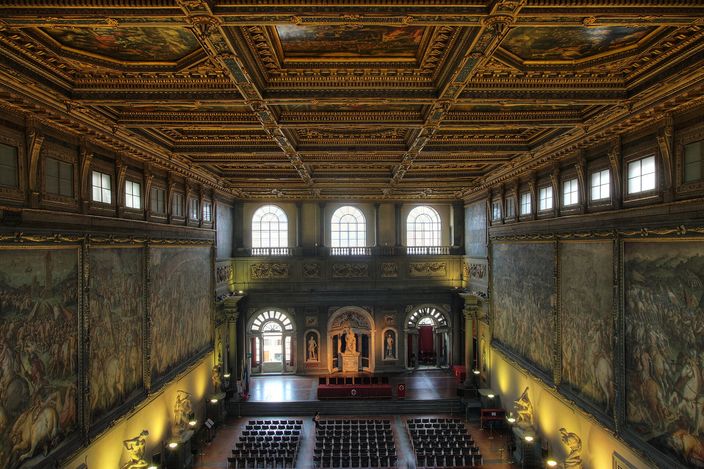 The Salone dei Cinquecento in Palazzo Vecchio seen from a balcony at one end: a vast hall with huge battle frescoes filling both long walls, a deep gilded coffered ceiling of painted panels overhead, rows of chairs set out on the floor and lit statues along the base of the walls