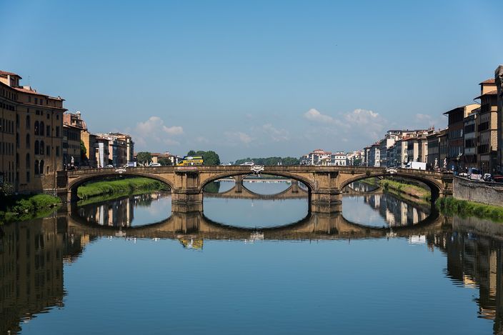 Ponte Santa Trinita seen along the Arno from the Ponte Vecchio on a still morning: three very shallow stone arches carried on two pointed cutwaters, their curves completed into full circles by the reflection in the flat water, with ochre houses and green embankments running away on both banks under a clear sky