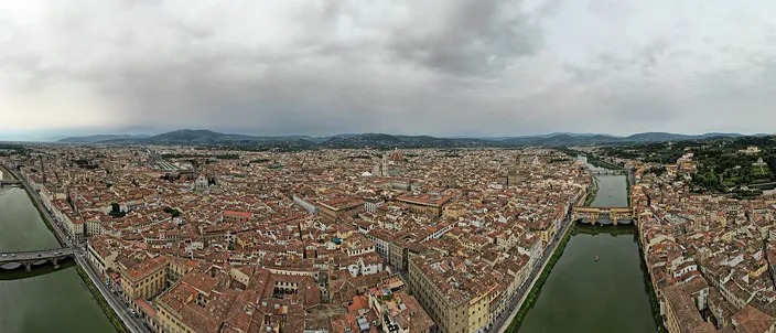 Florence from the air in flat daylight: the dense red-tiled roofs of the historic center filling the frame, the cathedral dome and bell tower standing above them left of center, the Arno curving away on the right past a bridge, and wooded hills along the horizon under heavy gray cloud