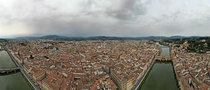 Florence from the air in flat daylight: the dense red-tiled roofs of the historic centre filling the frame, the cathedral dome and bell tower standing above them left of centre, the Arno curving away on the right past a bridge, and wooded hills along the horizon under heavy grey cloud