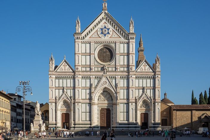 The marble facade of the basilica of Santa Croce in green and white geometric panels under a clear blue sky, with people crossing the open piazza in front of it