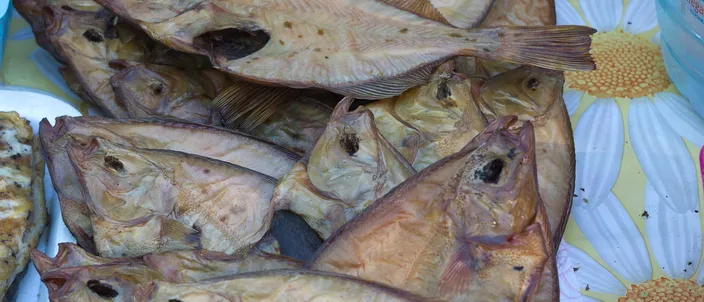 Whole hot-smoked flounder heaped on a beach stall, skins bronzed unevenly from gold to dark brown, tails and fins fanned out over a flowered tablecloth.