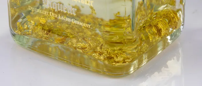 Close-up of the base of a Danziger Goldwasser bottle, with flakes of real gold leaf settled in the clear liqueur above the embossed 'Liqueur, Distillery Der Lachs Germany' lettering.