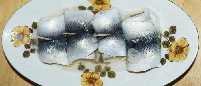 Oval floral-rimmed plate of four rolled pickled herring fillets (rolmopsy) held with wooden picks, with onion visible inside the rolls, on a wooden table.