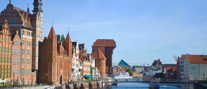 The Motława river in Gdańsk at midday, with the medieval brick Crane and Mariacka Gate on the Long Embankment, gabled red-brick houses along the quay and a bridge crossing the blue water.