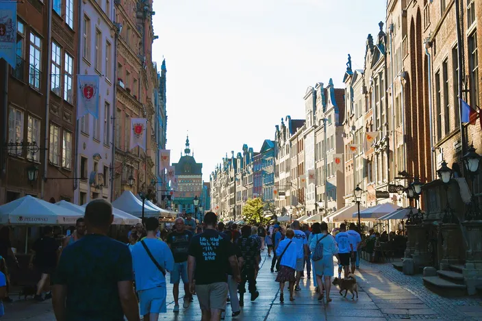 Long Street in Gdańsk on a summer day, crowded with walkers between tall gabled merchant houses hung with city flags, café umbrellas on both sides and a gate tower closing the view at the far end.
