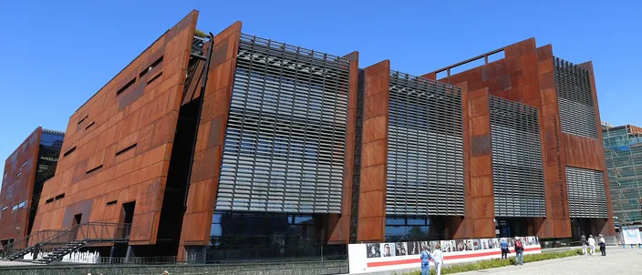 The European Solidarity Centre in Gdańsk under a clear sky: tall slanted panels of rust-colored steel alternating with louvred glass, a photo exhibition along the fence and a few people on the wide paved square in front.