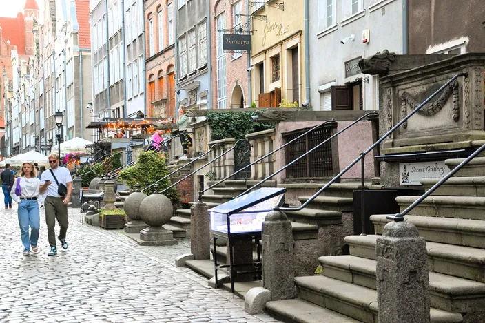 Mariacka Street in Gdańsk: a cobbled lane lined with raised stone terraces in front of each house, carved stone balustrades and steps, café tables and a few walkers between the tall gabled facades.