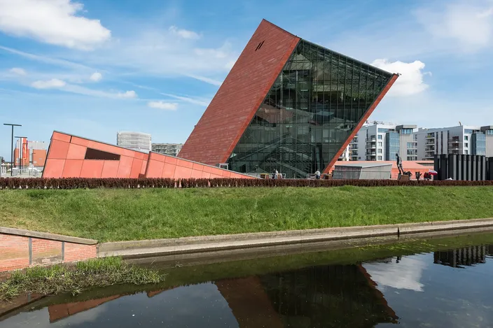 The Museum of the Second World War in Gdańsk, a dark red wedge-shaped building with a glazed front leaning out of the ground, seen across a canal with its reflection in the water under a blue sky.