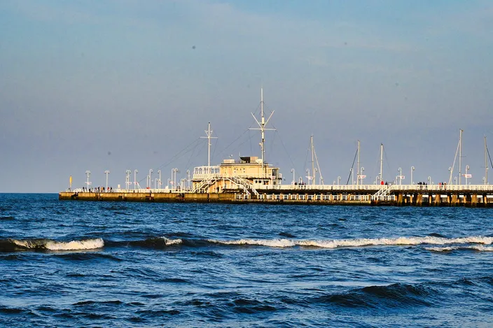 Sopot Pier seen from the beach, a long white wooden pier with a central pavilion and masts stretching across the blue Baltic, small waves breaking in the foreground.