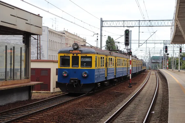 A blue-and-yellow SKM Trójmiasto electric suburban train pulls into Gdańsk Wrzeszcz station along a curved platform under overhead wires and signal gantries.