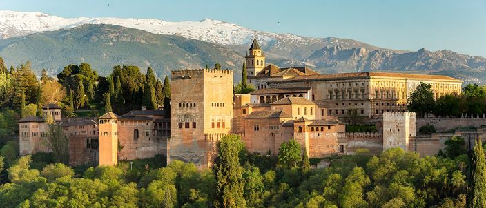 The Alhambra's ochre palace-fortress and towers rising above dense green woodland at golden hour, with the snow-capped Sierra Nevada behind, seen from the Mirador de San Nicolás.