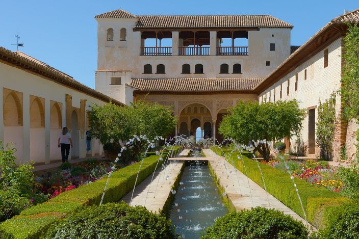The Patio de la Acequia in the Generalife: a long narrow courtyard with a water channel down its centre and two rows of jets arching across it, flower beds and clipped hedges either side, arcaded white galleries along both walls and a pavilion with an open loggia at the far end.