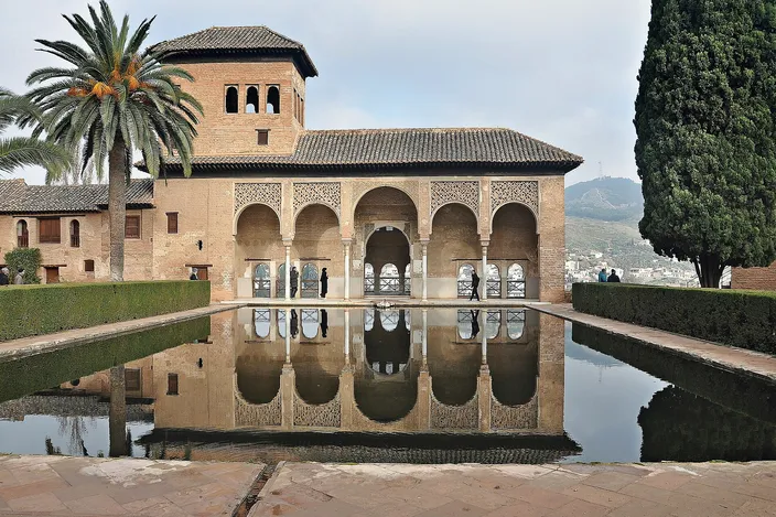 The Partal in the Alhambra: a five-arched brick portico with a square tower rising behind it, standing at the edge of a long still pool that reflects the whole building upside down, a palm tree at the left and a tall cypress at the right.