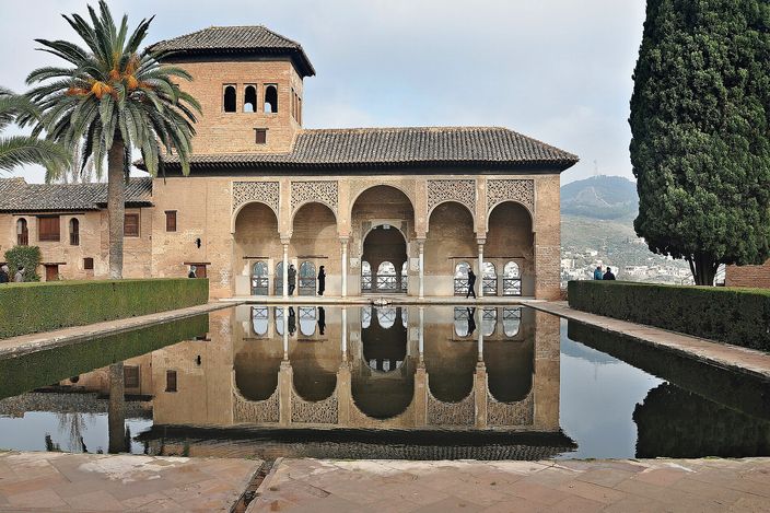 The Partal in the Alhambra: a five-arched brick portico with a square tower rising behind it, standing at the edge of a long still pool that reflects the whole building upside down, a palm tree at the left and a tall cypress at the right.