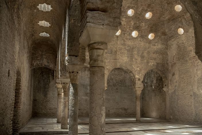 The central hall of El Bañuelo: three brick barrel vaults on stone columns whose capitals do not match, with star-shaped and octagonal holes cut through the vaults letting in daylight, and horseshoe-arched openings along the far wall.