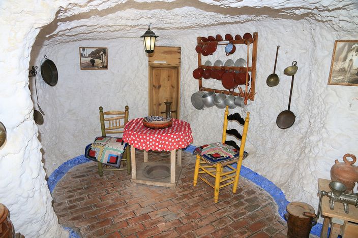 A furnished room cut into rock inside the Sacromonte cave museum: whitewashed walls and a low curved ceiling, a round table under a red-and-white checked cloth, two painted wooden chairs, a rack of copper pans and pots hanging on the wall, and a brick floor.