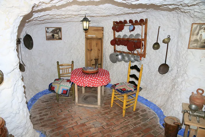 A furnished room cut into rock inside the Sacromonte cave museum: whitewashed walls and a low curved ceiling, a round table under a red-and-white checked cloth, two painted wooden chairs, a rack of copper pans and pots hanging on the wall, and a brick floor.