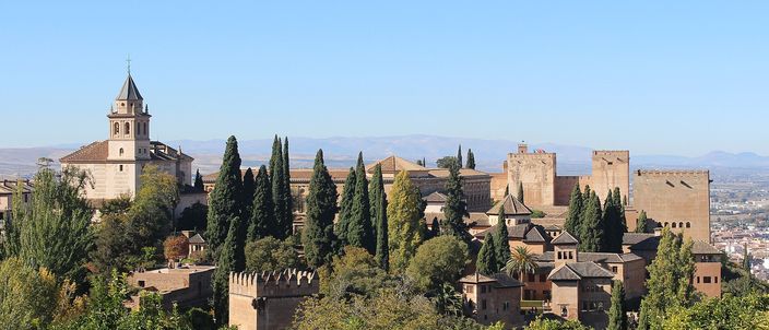 The Alhambra seen from the Generalife across a wooded valley: a church bell tower at the left, a row of dark cypresses through the middle, the ochre palace roofs and square battlemented towers of the fortress along the ridge, and the flat city and plain fading away at the right.