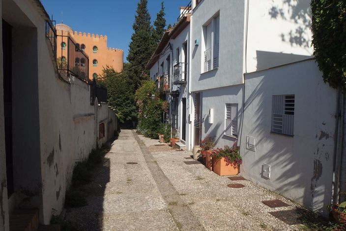 A narrow whitewashed lane paved with cobbles in Granada's Realejo quarter, houses on the right with potted plants, and an orange Moorish-style tower rising above trees at the end under a clear blue sky