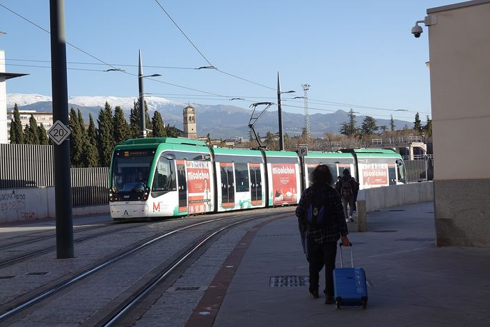 A green-and-white CAF Urbos tram at a Granada Metropolitano stop, a traveller with a wheeled suitcase walking towards it, snow-capped Sierra Nevada peaks and a church tower behind.