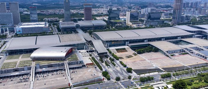 Aerial view over the Canton Fair exhibition complex in Pazhou: rows of low grey exhibition halls stretching across a whole riverside district, a red curved pavilion at the left, wide empty forecourts and a boulevard, with office towers and the hazy city behind
