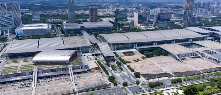 Aerial view over the Canton Fair exhibition complex in Pazhou: rows of low gray exhibition halls stretching across a whole riverside district, a red curved pavilion at the left, wide empty forecourts and a boulevard, with office towers and the hazy city behind