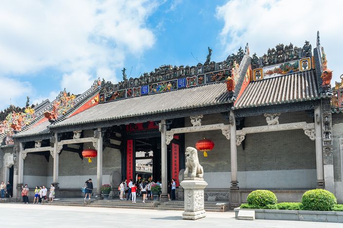 The front elevation of a Chinese ancestral hall in grey brick and granite, its long roof ridges crowded with brightly painted ceramic figures, red lanterns hung between the carved timber brackets and a stone lion on a plinth in the forecourt.