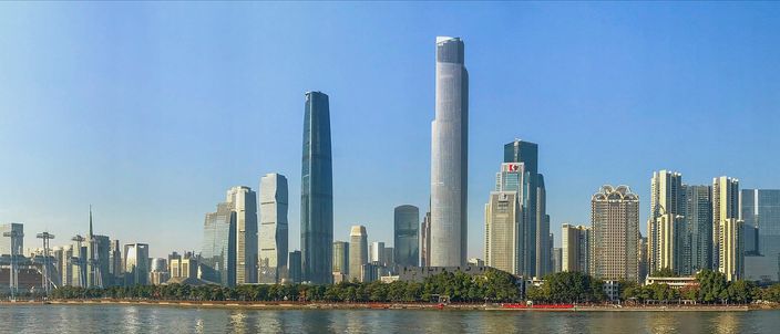 A wide daytime view across the Pearl River to a dense cluster of glass towers, a tall pale supertall rising at the centre and a tapered one beside it, with a green bank and a low red-roofed pier along the water's edge.
