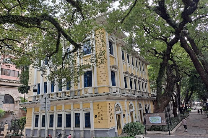 A two-storey cream and yellow colonial building with an arcaded ground floor and shuttered windows, standing on a quiet street beneath the enormous spreading branches of banyan trees.
