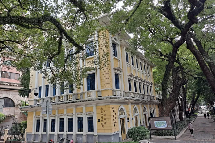 A two-story cream and yellow colonial building with an arcaded ground floor and shuttered windows, standing on a quiet street beneath the enormous spreading branches of banyan trees.