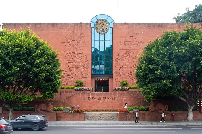 The symmetrical red sandstone front of a museum, carved with large abstract reliefs either side of a tall glazed slot and a bronze medallion, with steps, planting and a road in front of it.