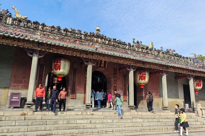 The front of a Chinese temple hall seen from its steps, the whole roof ridge packed with small painted ceramic figures against a blue sky, two large red lanterns hanging between the columns and visitors going in and out of the open doorway.