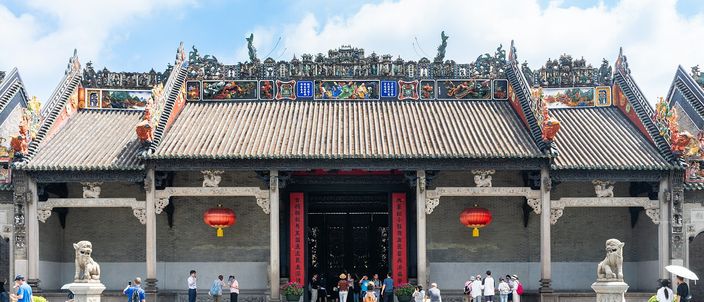The wide grey-brick and granite front of a Chinese ancestral hall under a blue sky, its long roof ridges crowded with brightly painted ceramic figures, red lanterns hung beneath the carved timber eaves and a stone lion on a plinth at each end of the forecourt.