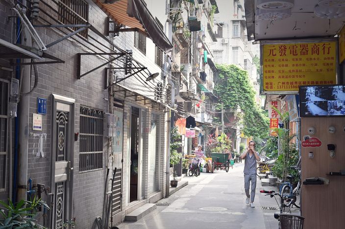 A narrow lane in Guangzhou's old centre: grey brick house fronts with barred windows on the left, laundry poles jutting from upper floors, potted plants and a handcart further down, and a man walking towards the camera in the shade