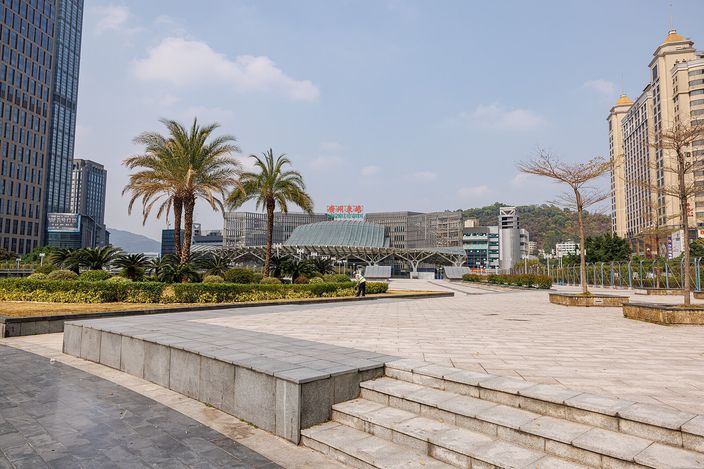 The plaza in front of Guangzhou East railway station on a clear day: broad pale paving and low steps in the foreground, palm trees and clipped hedges, the station's long glass-and-steel terminal building in the middle distance, office towers to the left and green hills behind