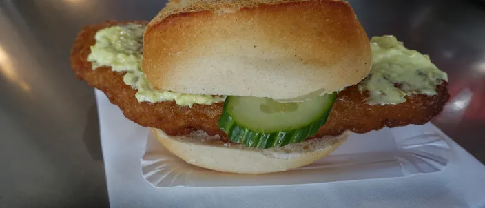 A Fischbrötchen in a crusty white roll filled with a piece of fried fish, a slice of pickled gherkin and remoulade, served on paper
