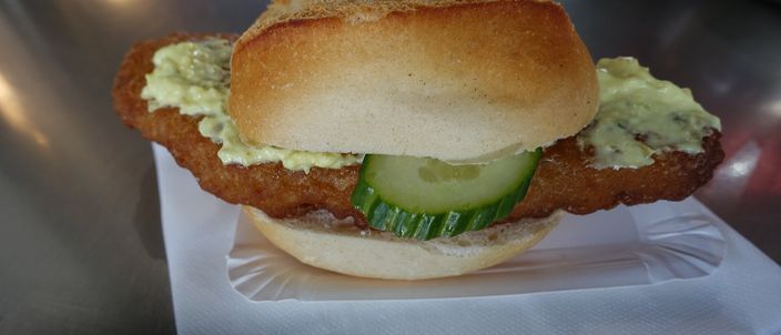 A Fischbrötchen in a crusty white roll filled with a piece of fried fish, a slice of pickled gherkin and remoulade, served on paper