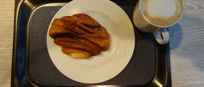 A single Franzbrötchen on a white plate beside a cappuccino, showing the flattened, caramelized cinnamon pastry