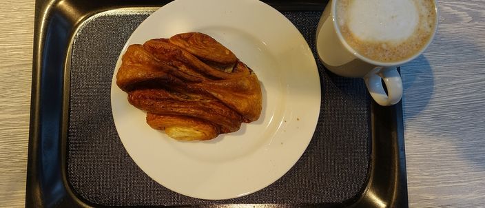 A single Franzbrötchen on a white plate beside a cappuccino, showing the flattened, caramelised cinnamon pastry