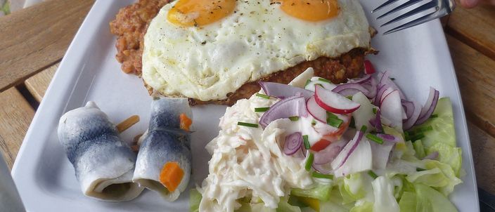 A plate of Labskaus: a pink-tinged beef, potato and beetroot hash topped with two fried eggs, with rolled pickled herring and an onion salad on the side
