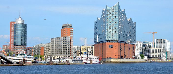 The Elbphilharmonie's wave-shaped glass roof rising above its red-brick base on the Elbe waterfront, flanked by HafenCity towers and moored ships under a clear sky.