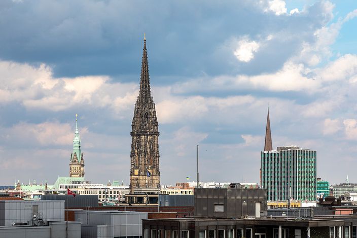 The blackened stone spire of the ruined St. Nikolai church rising above the flat modern rooftops of central Hamburg, with the green copper tower of the town hall to the left and a slender brown church spire beside a green glass office block to the right, under a sky of broken cloud.